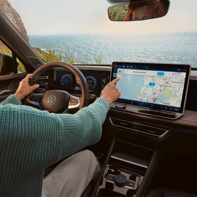Eine Person navigiert auf einem Touchscreen in einem Volkswagen, während sie entlang einer Küstenstraße fährt.