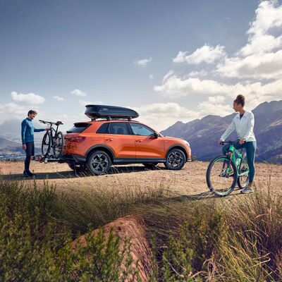Ein orangefarbenes Auto mit Fahrradträger im Gebirge, zwei Personen mit Fahrrädern. Audi Zentrum Passau.