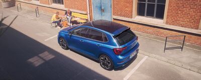 Ein blauer Škoda parkt vor einem Backsteingebäude, während zwei Personen auf einer Bank sitzen und sich unterhalten.