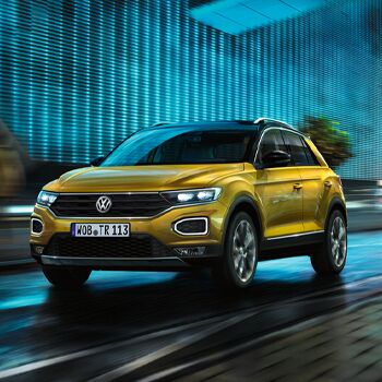 Ein gelber Volkswagen T-Roc fährt dynamisch bei Nacht durch eine beleuchtete Stadtstraße, präsentiert von Auto Röhr.