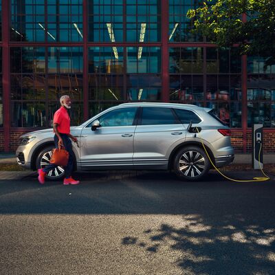 Ein Mann geht an einem silbernen SUV vorbei, der an einer Ladestation angeschlossen ist. | Auto Röhr