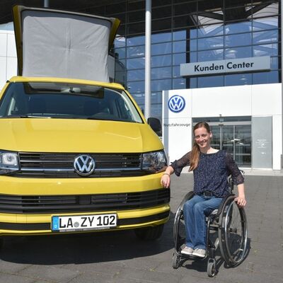 Eine Frau im Rollstuhl lächelt vor einem gelben Volkswagen vor dem Kunden Center von Volkswagen Zentrum Passau.