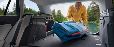 Ein Mann lädt eine blaue Tasche in den Kofferraum eines neuen Volkswagen Passat an einem See. Volkswagen Zentrum Passau.