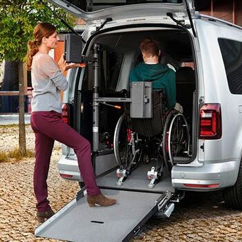 Eine Frau hilft einem Mann im Rollstuhl, in einen silbernen Van mit Rampe einzusteigen