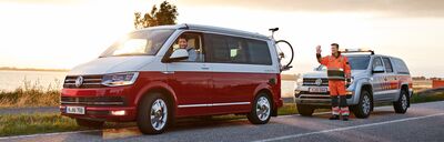 Ein roter Volkswagen-Bus hält auf der Straße, während ein Pannenhelfer freundlich neben einem Servicefahrzeug steht.