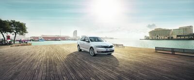 Škoda Rapid steht auf einer hölzernen Plattform am Wasser mit modernen Gebäuden im Hintergrund. Škoda Auto Röhr.