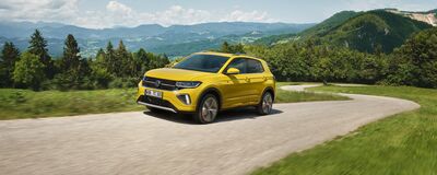 Gelber Volkswagen T-Cross fährt auf einer kurvigen Landstraße vor einer malerischen Berglandschaft. Škoda Auto Röhr.
