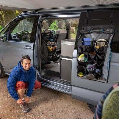 Ein Mann kniet lächelnd neben einem grauen Camper mit geöffneter Tür und organisiertem Innenraum. | Auto Röhr.