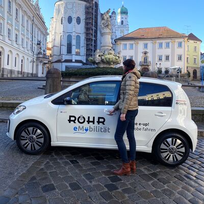 Frau öffnet die Tür eines weißen Volkswagen e-up! mit Auto Röhr Werbung in einer historischen Stadtumgebung.