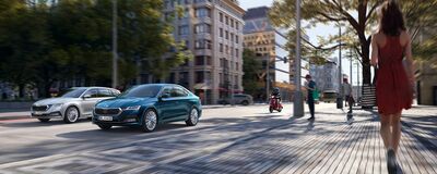 Zwei Škoda Octavia fahren durch eine belebte Stadtstraße. Ein Fußgänger und ein Roller sind im Hintergrund. Škoda Auto Röhr.