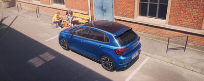 Ein blauer Volkswagen parkt vor einer Ziegelmauer, während zwei Personen entspannt am Tisch sitzen | © Erich Röhr GmbH & Co. KG.