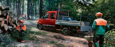 Ein roter VW Crafter Pritschenwagen steht im Wald, während Arbeiter Holzstämme bearbeiten. Škoda Auto Röhr.