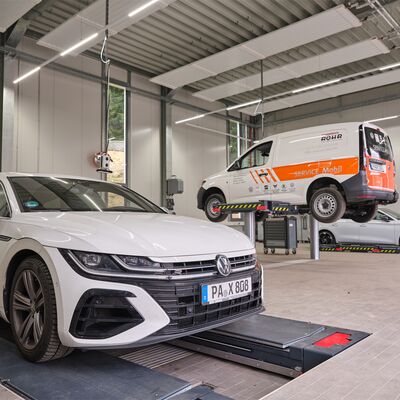 VW-Werkstatt mit weißem Auto auf Hebebühne und Servicefahrzeug von Auto Röhr im Hintergrund.
