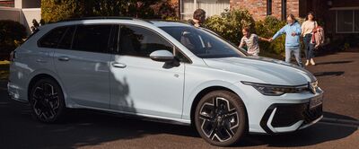 Ein weißer Volkswagen Kombi parkt vor einem Haus, während eine Familie im Hintergrund steht. Volkswagen Zentrum Passau.