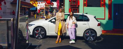 Zwei Frauen lehnen an einem weißen Volkswagen Golf GTE vor bunten Gebäuden. | © Auto Röhr