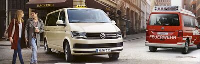 Ein weißes VW-Taxi und ein rotes Feuerwehrfahrzeug auf einer Stadtstraße. Passanten laufen vorbei. Volkswagen Zentrum Passau.