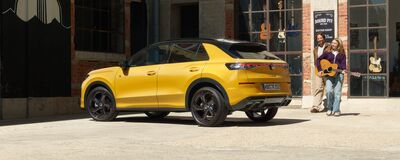 Ein gelbes Auto steht vor einem Gitarrenladen, während zwei Personen mit einer Gitarre plaudern.