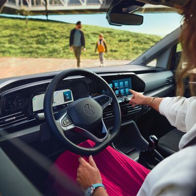 Frau im Volkswagen, navigiert Touchscreen. Im Hintergrund gehen Mann und Kind. | Škoda Auto Röhr.