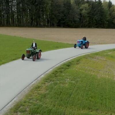 Vier Traktoren und ein Roller fahren auf einer ländlichen Straße durch grüne Felder, inspiriert von Bulldog und Rolletta.