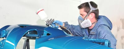 Ein Lackierer trägt blaue Farbe auf ein Autoteil auf. Schutzmaske und Spritzpistole sind im Einsatz. Auto Röhr.