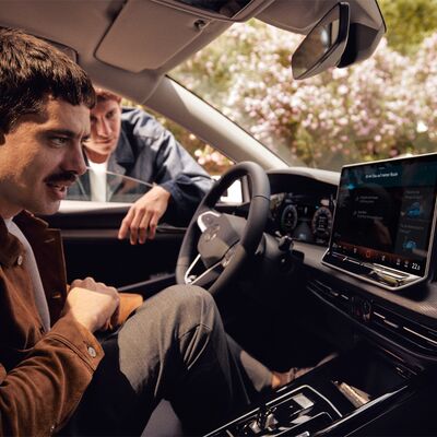 Mann sitzt in einem Auto und betrachtet das Infotainmentsystem, während ein anderer Mann von außen zuschaut. | Škoda Auto Röhr.