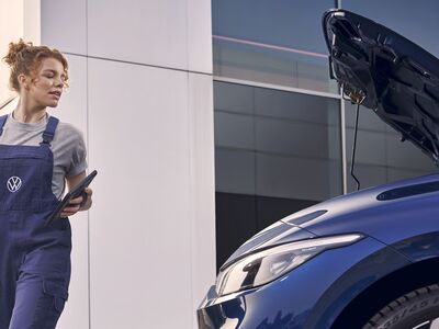 Mechanikerin inspiziert ein Auto mit geöffnetem Motorhaube vor einem modernen Gebäude. Volkswagen Zentrum Passau.