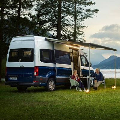 Ein Paar sitzt vor einem Volkswagen Grand California Camper am See, umgeben von Bergen. | © Auto Röhr