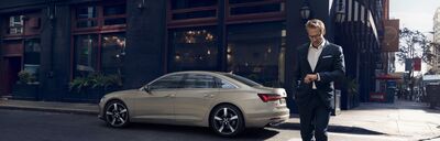 Ein Mann im Anzug geht an einem geparkten Audi vor einem modernen Gebäude vorbei. | Audi Zentrum Passau.