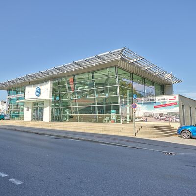 Das Volkswagen Zentrum Passau zeigt moderne Glasarchitektur und ein blaues Auto Röhr parkt davor.