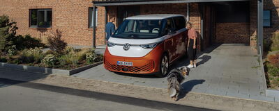 Eine Frau und ein Mann stehen neben einem Volkswagen ID. Buzz im Carport, während ein Hund um sie herumlaufen.