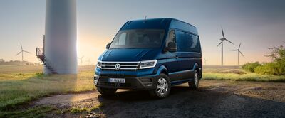 Blauer VW Crafter auf einem Feldweg bei Sonnenaufgang mit Windrädern im Hintergrund. | Škoda Auto Röhr