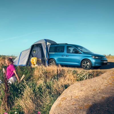 Ein blaues Auto mit angebautem Zelt auf einem Feld, ein Kind läuft vorbei. Škoda Auto Röhr.