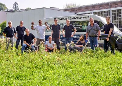 Eine Gruppe von Mitarbeitern posiert fröhlich vor einem Lieferwagen von Auto Röhr auf einer blühenden Wiese.