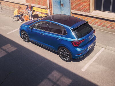 Ein blauer Volkswagen steht in einer sonnigen Straße, während zwei Personen auf einer Bank entspannt sind. | © Volkswagen