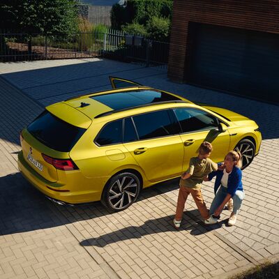 Gelber Volkswagen Kombi auf gepflasterter Auffahrt, neben einer Frau und einem Kind. Volkswagen Zentrum Passau.