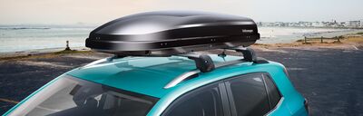 Ein türkisfarbenes Auto mit Dachbox steht am Meer, präsentiert von Volkswagen Zentrum Passau.