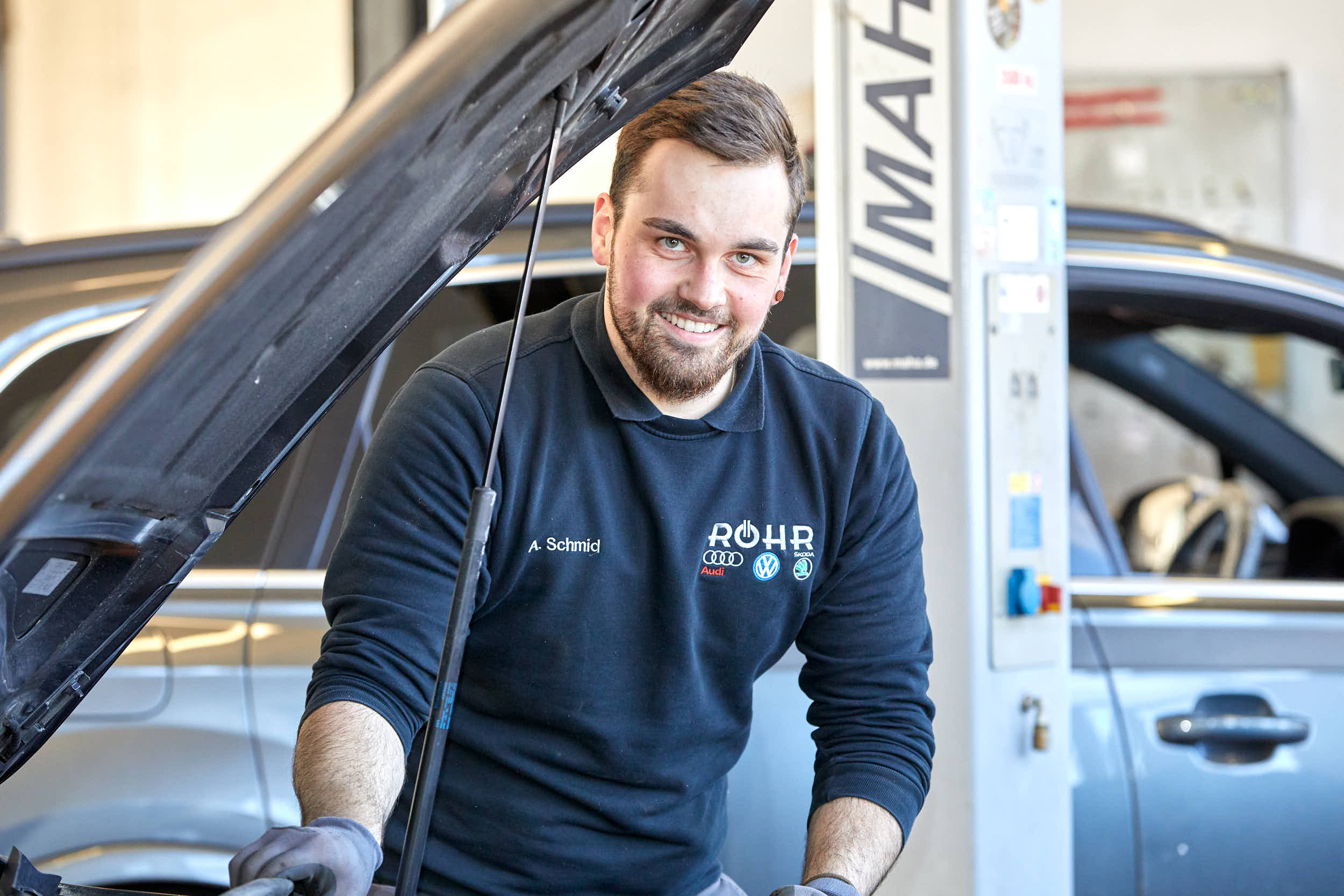 Mechaniker lächelt neben einem Auto mit geöffnetem Motorraum in der Werkstatt von Auto Röhr.