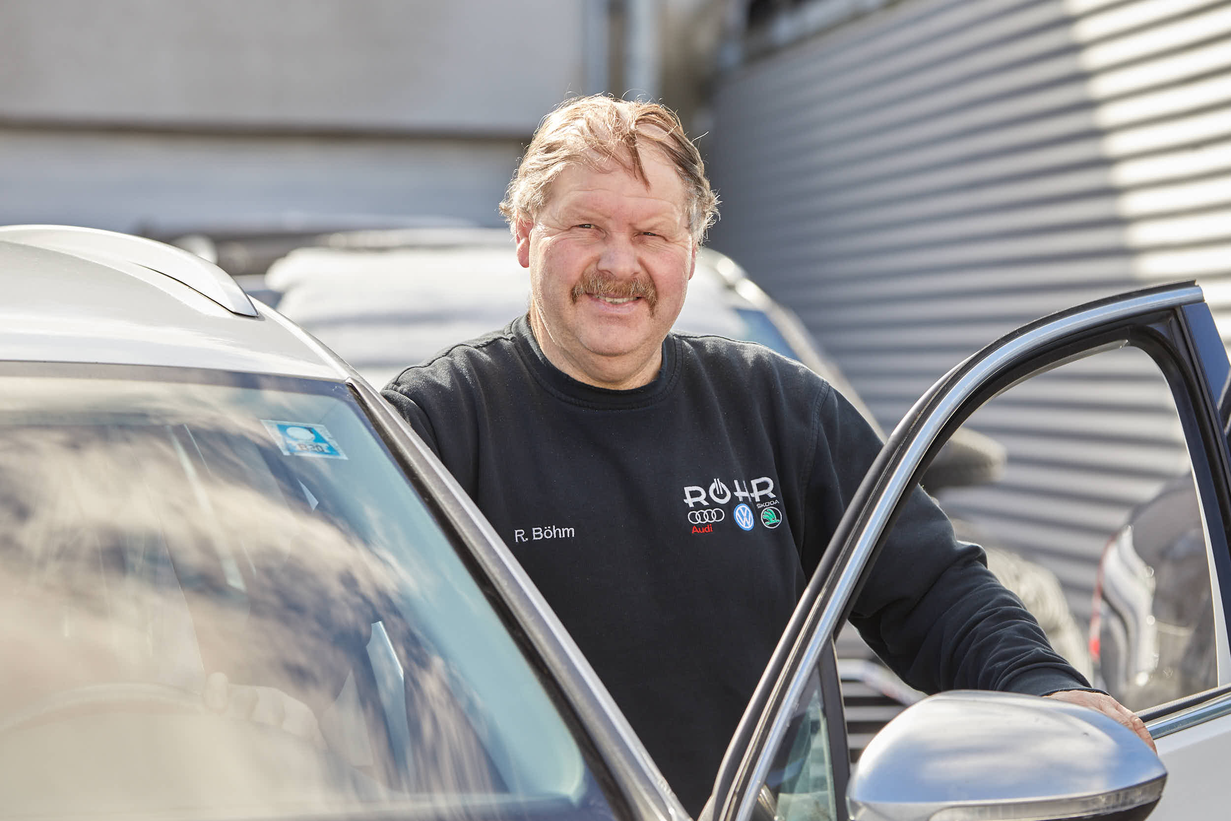 Ein Mann in einem Autohaus öffnet die Tür eines Autos. Er trägt ein Sweatshirt mit Logos von Auto Röhr.