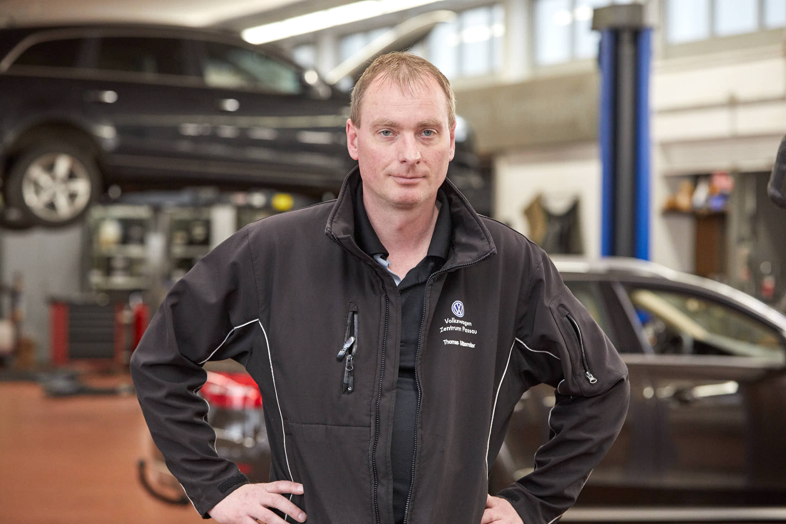 Ein Mann in Arbeitskleidung steht in einer Autowerkstatt, im Hintergrund ein angehobenes Fahrzeug. Volkswagen Zentrum Passau.