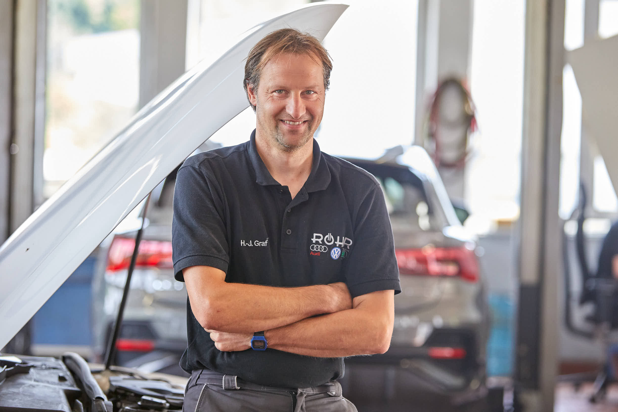 Mechaniker im Audi Zentrum Passau steht lächelnd vor einem Auto mit geöffnetem Motorraum in einer Werkstatt.